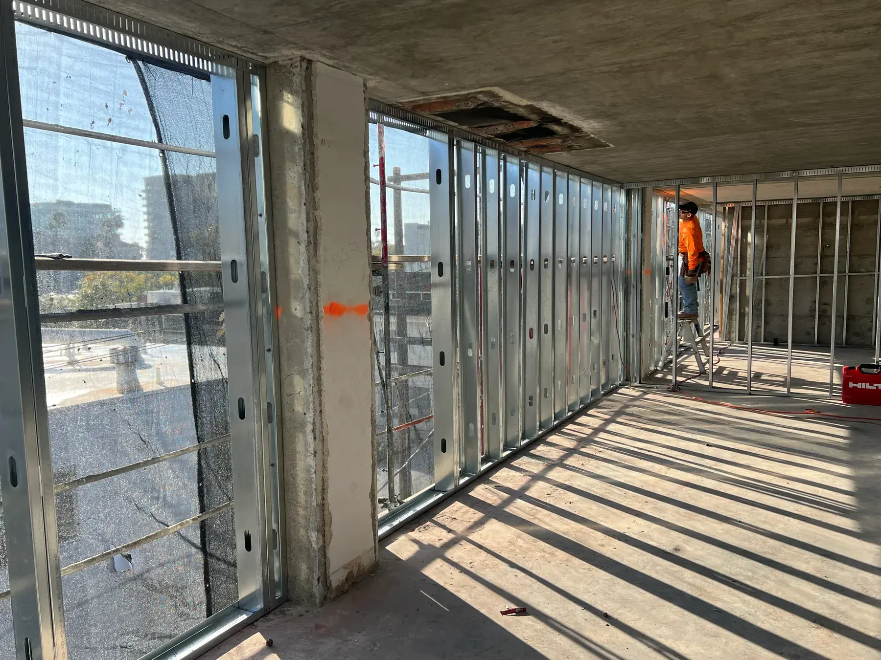 Steel stud exterior wall framing running the length of a concrete floor plate, with a crew member setting studs from a ladder